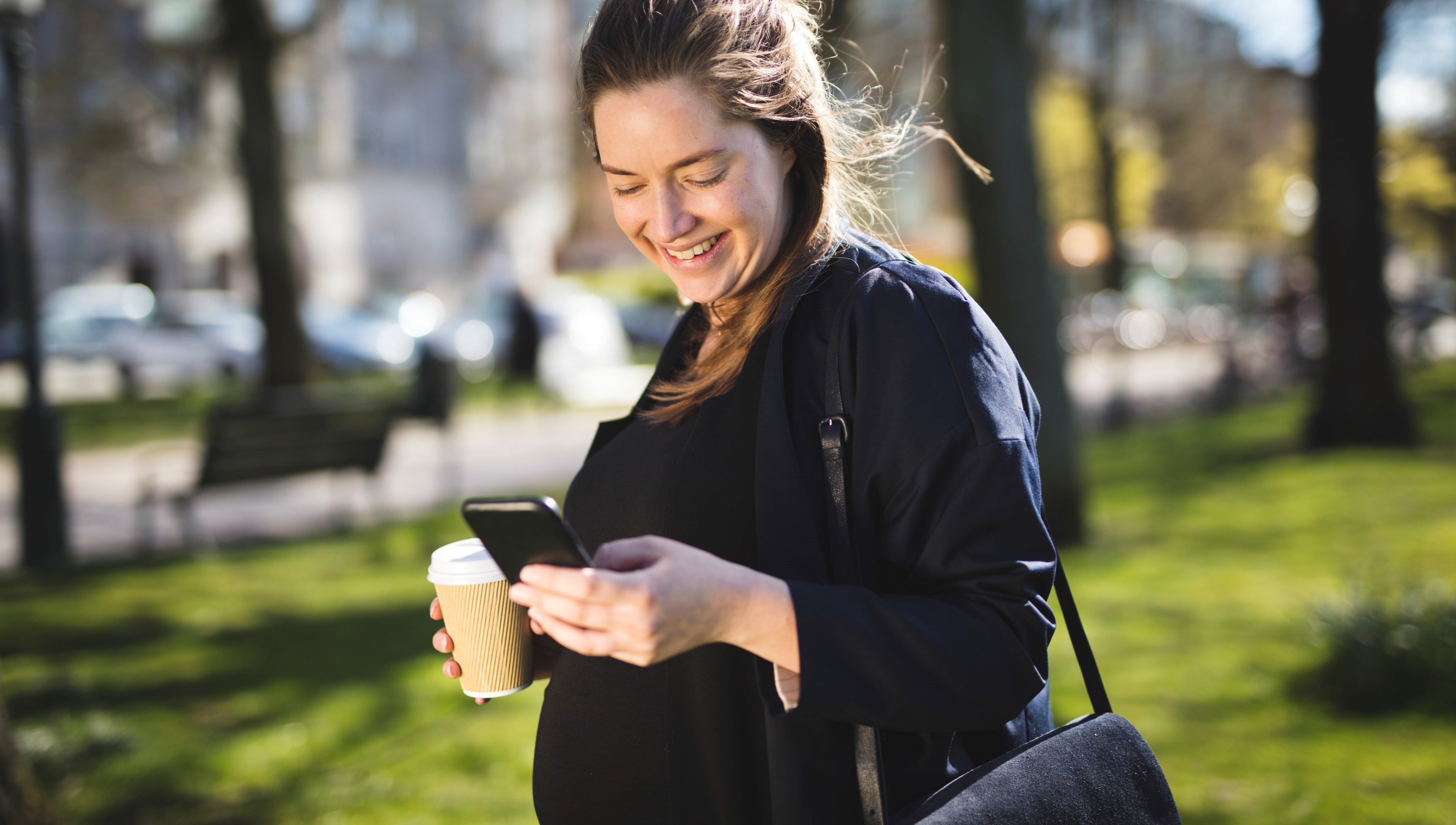 En gravid kvinna promenerar i naturen, hon ser på sin mobiltelefon och håller en mugg i handen.