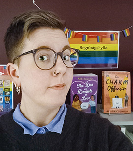 Emma Bladby har kortklippt brunt hår och bär glasögon med bruna bågar. Hon har en näspiercing och bär örhängen. Hon är klädd i en blå skjorta med en svart tröja över, bakom henne syns bibliotekets regnbågshylla.