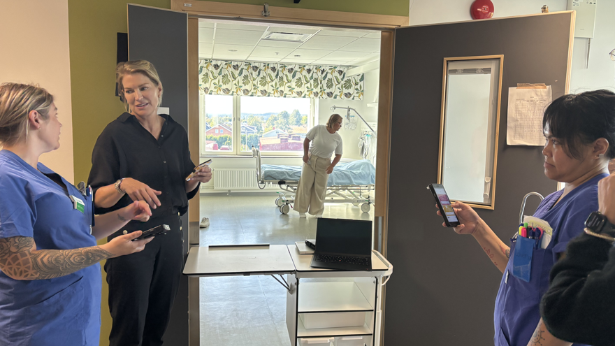 vårdpersonal i samspråk utanför ett vårdrum. I bakgrunden syns en person röra sig i vårdrummet bredvid en sjuksäng.