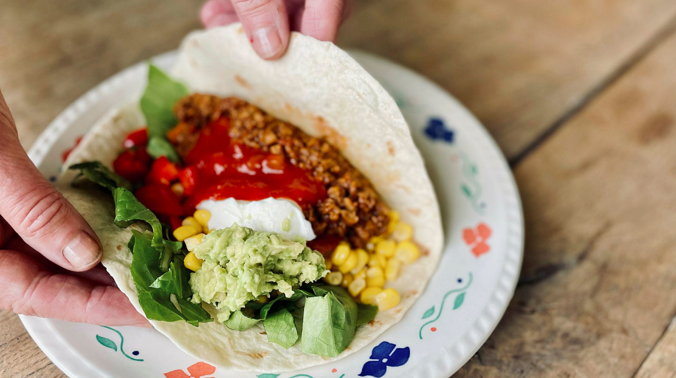 Händer som håller i ett tortillabröd fyllt med olika grönsaker och färs