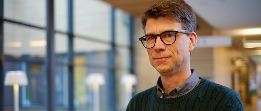 Man med mörkt hår och glasögon står i offentlig lokal.