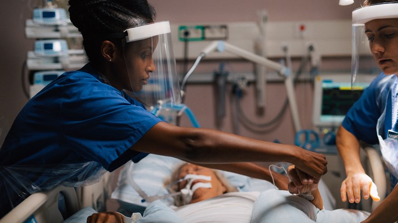 Två sjuksköterskor med visir tar hand om en patient i en sjukhussäng.