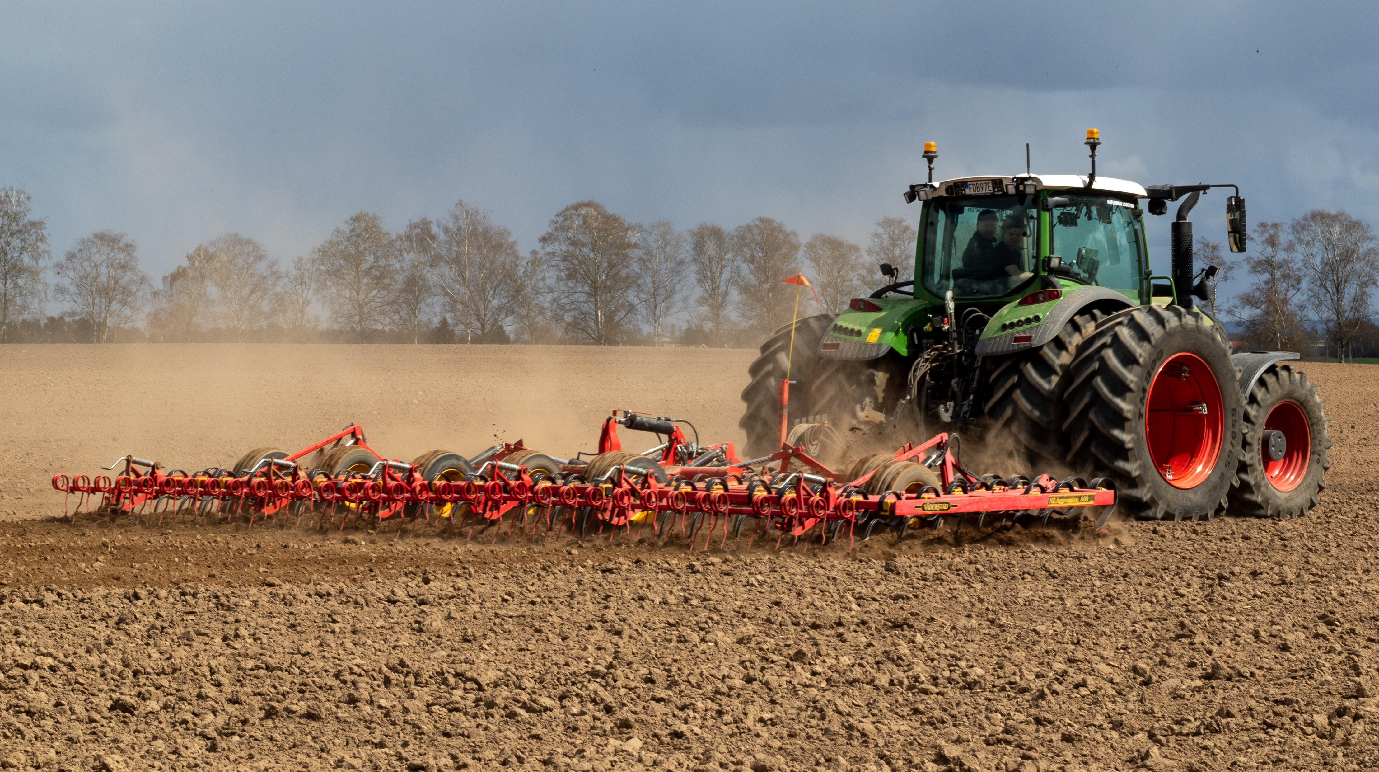 Traktor med harv på öppet fält med träd i bakgrunden