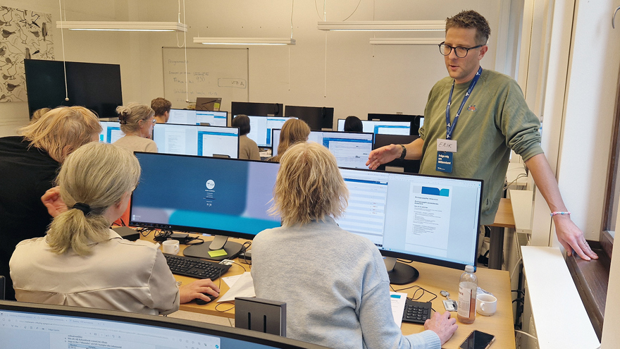 Ett utbildningsrum där deltagare sitter vid arbetsplatser med stora dator skärmar. En manlig utbildare står upp i rummet och förklarar något för en av deltagarna.