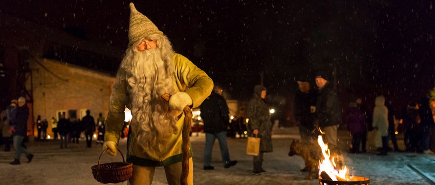 Tomte i grå kostym och långt skägg med en stav och en korg i ena handen