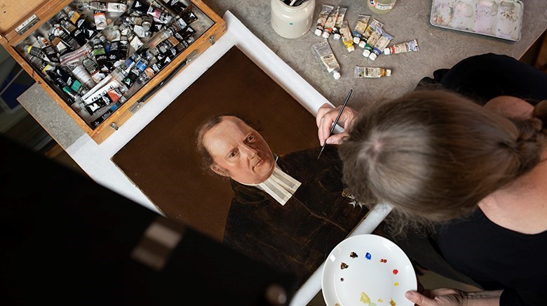 En porträttmålning ligger på ett bord. Intill finns en låda med mängder med färgtuber. Böjd över målningen står en person med pensel i handen och en palett med färg.