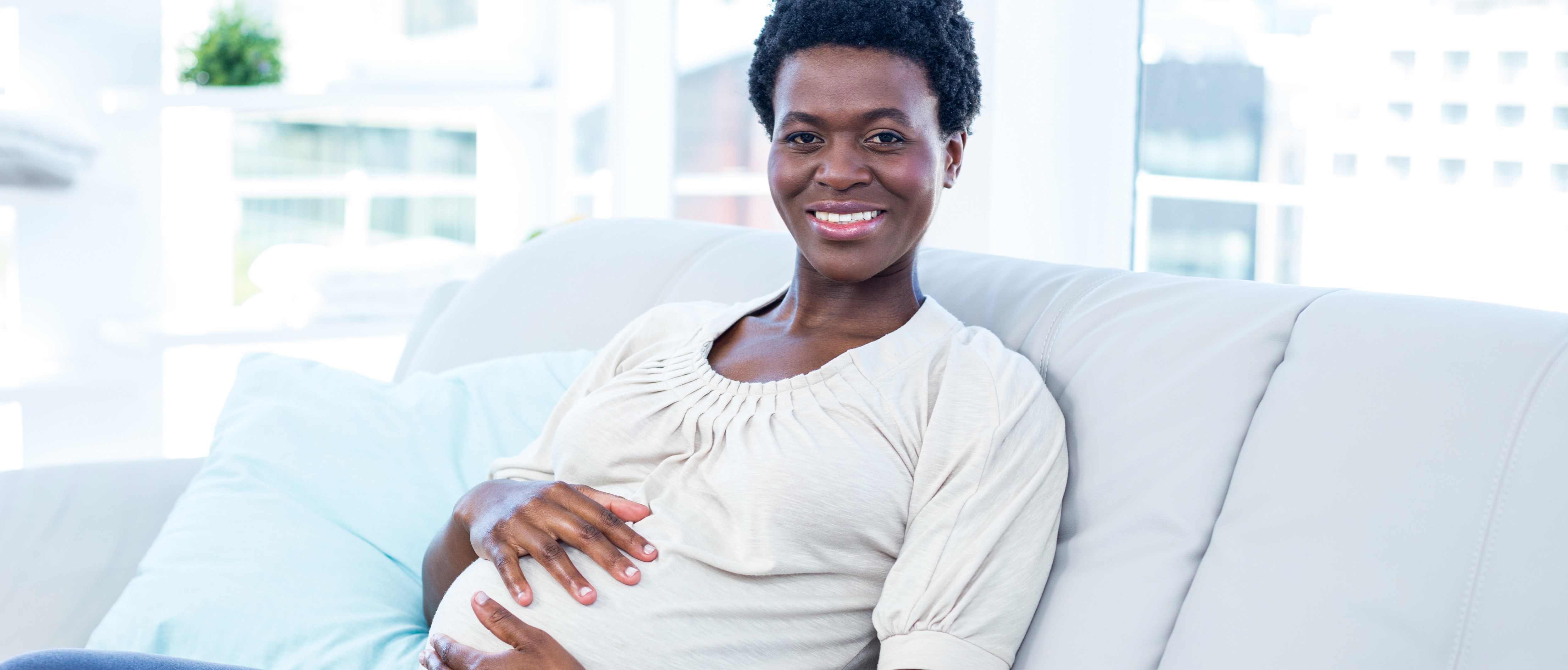 En gravid kvinna sitter i en soffa, ler och håller händerna på magen.