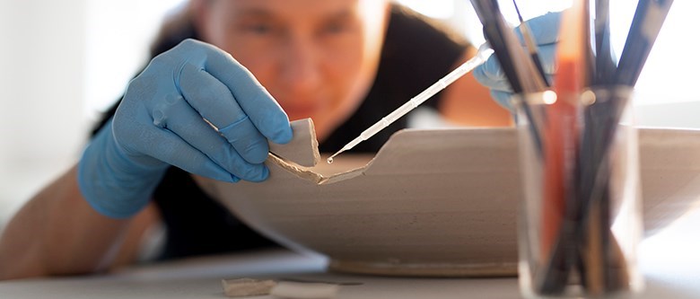 En kvinna limmar försiktigt en skärva på kanten av ett stort keramikfat. Hon har ljusblå latexhandskar på sig. I förgrunden står en burk med olika penslar i.
