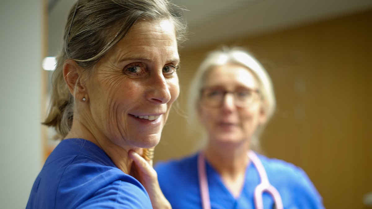 Kvinna i hälso- och sjukvårdskläder tittar in i kameran och ler.